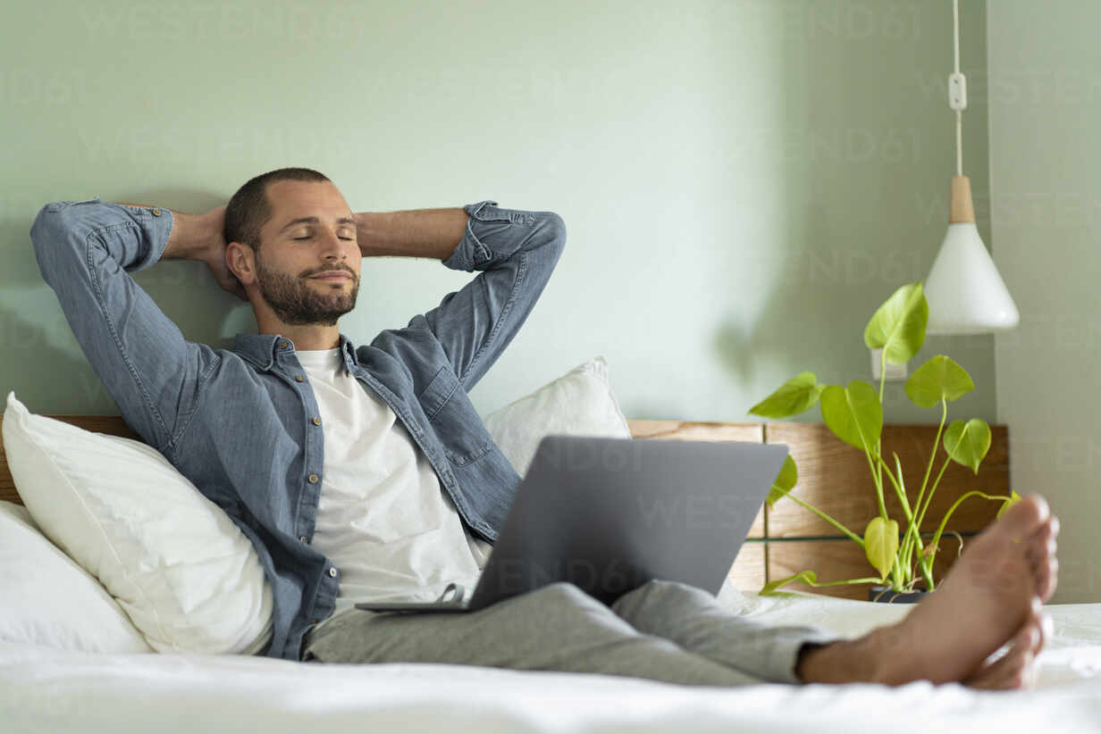 entspannter-mann-sitzt-mit-laptop-auf-dem-schoss-im-schlafzimmer-zu-hause-SBOF03218.jpg